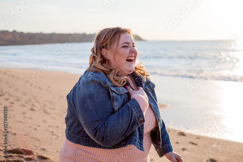 Beautiful overweight woman walking on the sandy beach. Plus size girl enjoy warmth sunset with romantic mood. Fat model dressed jeans jacket and pink knitted sweater photo