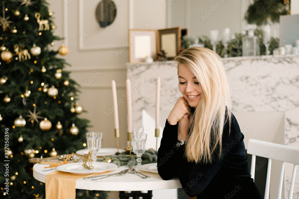 Christmas portrait of a smiling blonde girl in a black dress on the background of Christmas decor at a table in an elegant interior. A woman is preparing to celebrate Christmas and New year Christmas portrait of a smiling blonde girl in a black dress on the background of Christmas decor at a table in an elegant interior. A woman is preparing to celebrate Christmas and New year