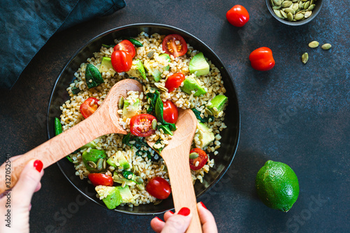 Hands mixing healthy salad with bulgur, avocado, spinach and cherry tomatoes. Top view. photo
