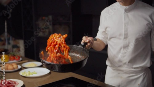 Panning tilt down medium shot of unrecognizable young chef tossing pasta with tomato sauce in pan in slow motion while cooking in restaurant kitchen