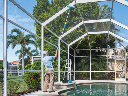 Handyman cleaning outdoor pool cage enclosure with pole brush. Screened swimming pool lanai maintenance and screen repair.