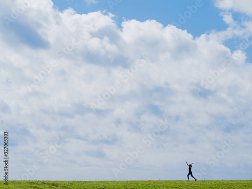 Sporty girl is full of joy and running on a green field with her hands up high with a blue sky with white clouds on the background. 