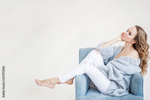 Attractive European girl with light brown hair in pastel clothes and beige shoes on a heel poses sitting on a light blue chair. Studio shooting. Light background.