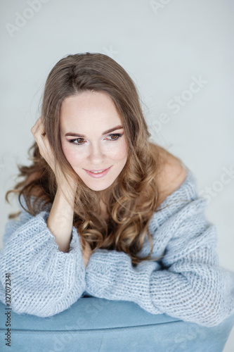 Close-up portrait of attractive young European smiling girl with light brown hair in light blue clothes. She poses sitting on a light blue chair. Studio shooting.  Light background.