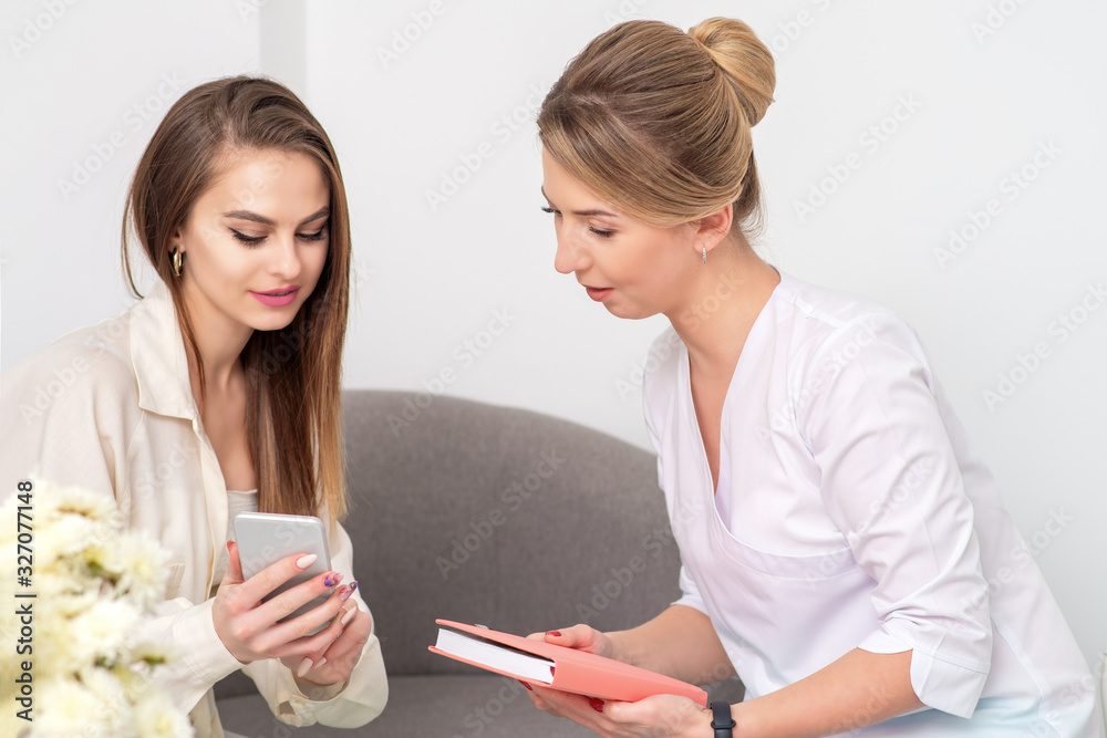 Cosmetologist talking with young woman client in bathrobe looking in smartphone of client. Cosmetologist talking with young woman client in bathrobe looking in smartphone of client.
