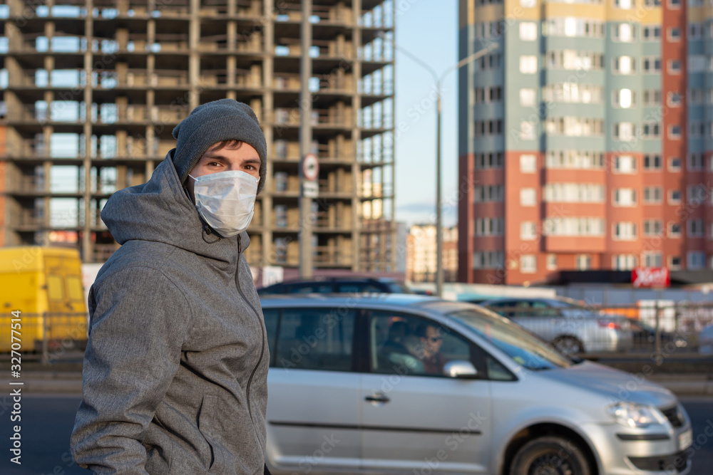 a guy is standing in the city near the road. Around unfinished apartments, traveling cars. Wearing a protective medical mask on his face. Danger of walking their viruses, coronavirus and diseases.