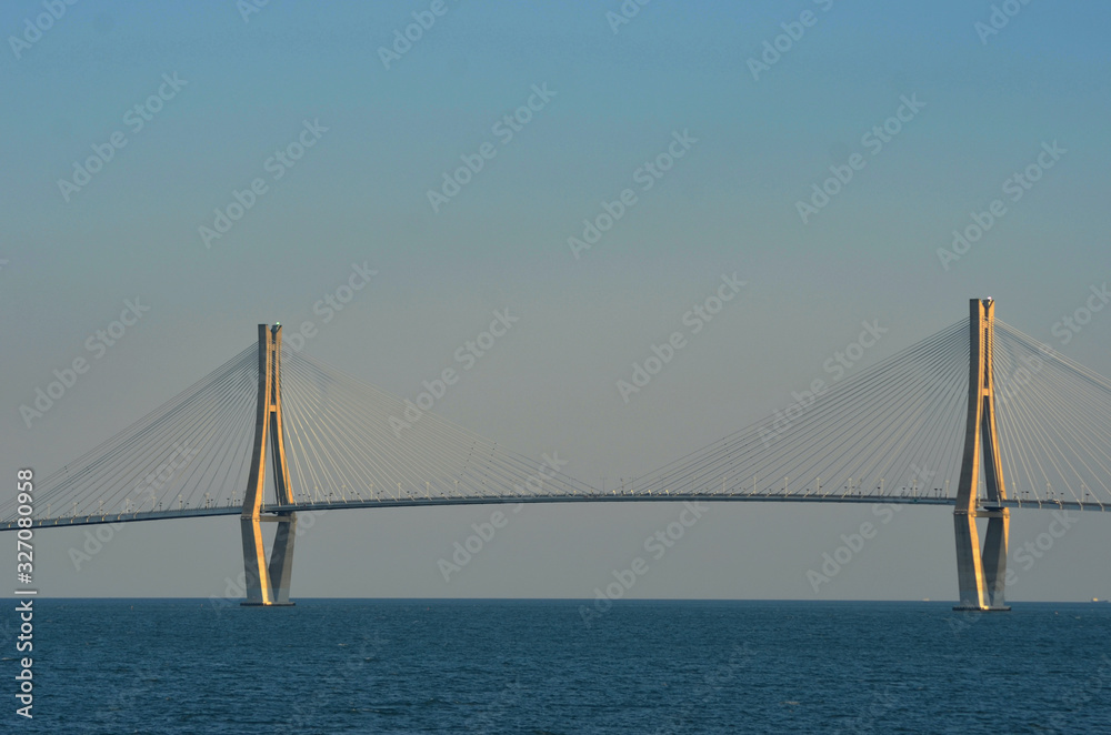 Fototapeta premium Two concrete pylons with radial supportive wires form a feature of a suspension bridge crossing a harbour. two tankers can be seen on the horizon. The sky is blue, with a haze near the horizon.
