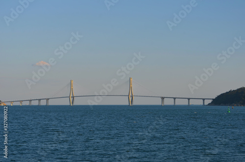 A bridge arches over a bay from an island to the mainland. Two pylons, with radiating supportive metal struts, rise above the bridge to form an architectural feature. The sky is blue with clouds.