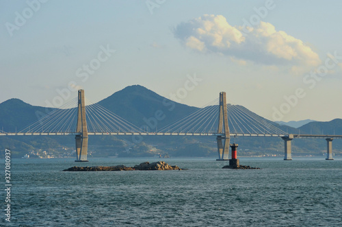 A bridge with two feature pylons stetches over a bay. Two rocky islands are in the foreground, one with a red channel marker. Mountains are in the background, with buildings on the foreshore. The sky 
