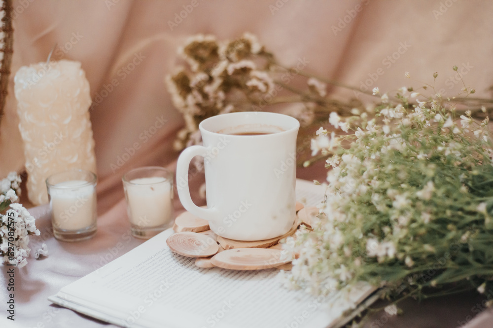 A cup of coffee on a stand surrounded by flowers and candles of different sizes and shapes A cup of coffee on a stand surrounded by flowers and candles of different sizes and shapes