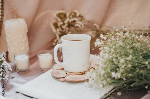 A cup of coffee on a stand surrounded by flowers and candles of different sizes and shapes