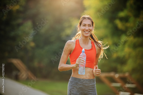 Fototapeta Naklejka Na Ścianę i Meble -  Young smiling positive attractive sportswoman with long hair holding bottle of water and running in public park in the morning.