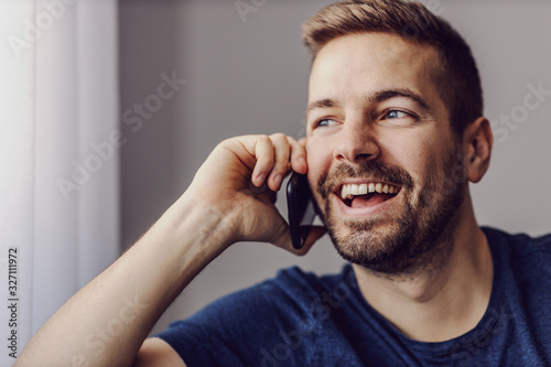 Close up of young handsome smiling unshaven man having phone conversation with friend while looking trough window.