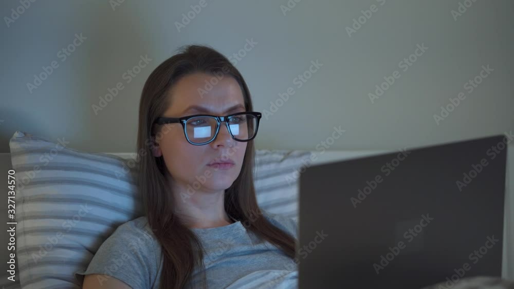 Woman in glasses working on laptop while lying in bed at night. Mobile addict or insomnia concept. Woman in glasses working on laptop while lying in bed at night. Mobile addict or insomnia concept.