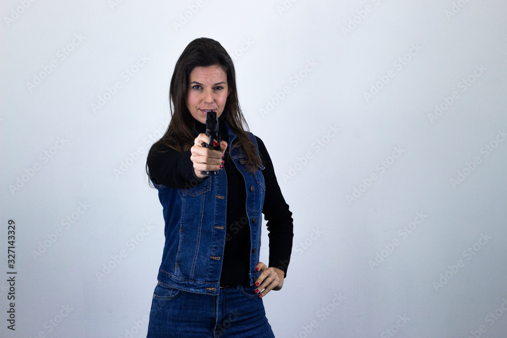 Portrait of fashionable middle age woman modelgun pointing with fore finger the object, isolated on gray background studio shotblack sweater, denim jacket, jeans, dark air. Place for your text in copy