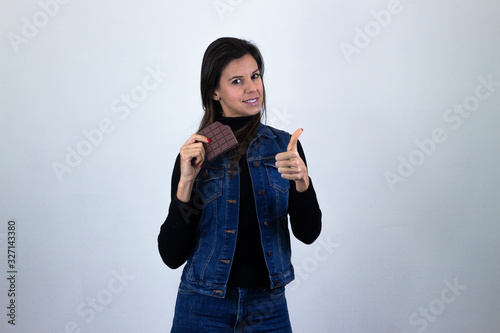 pretty  middle age woman model with chocolate showing thumbs up gesture, isolated on gray background studio shotblack sweater, denim jacket, jeans. Place for your text in copy space. photo