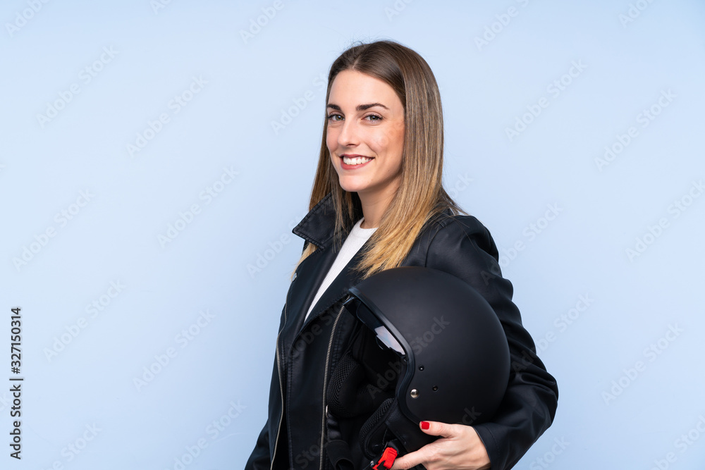 Young blonde woman with a motorcycle helmet over isolated blue background smiling a lot Young blonde woman with a motorcycle helmet over isolated blue background smiling a lot