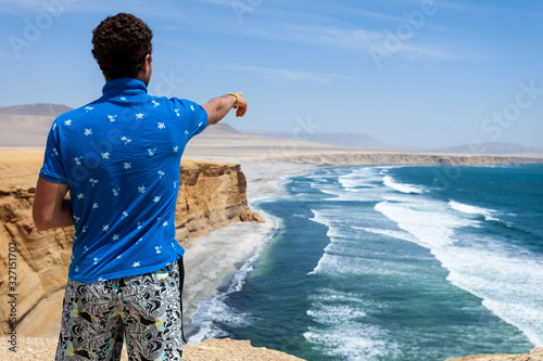 A man in a blue T-shirt stands on top of a cliff and points towards the ocean photo