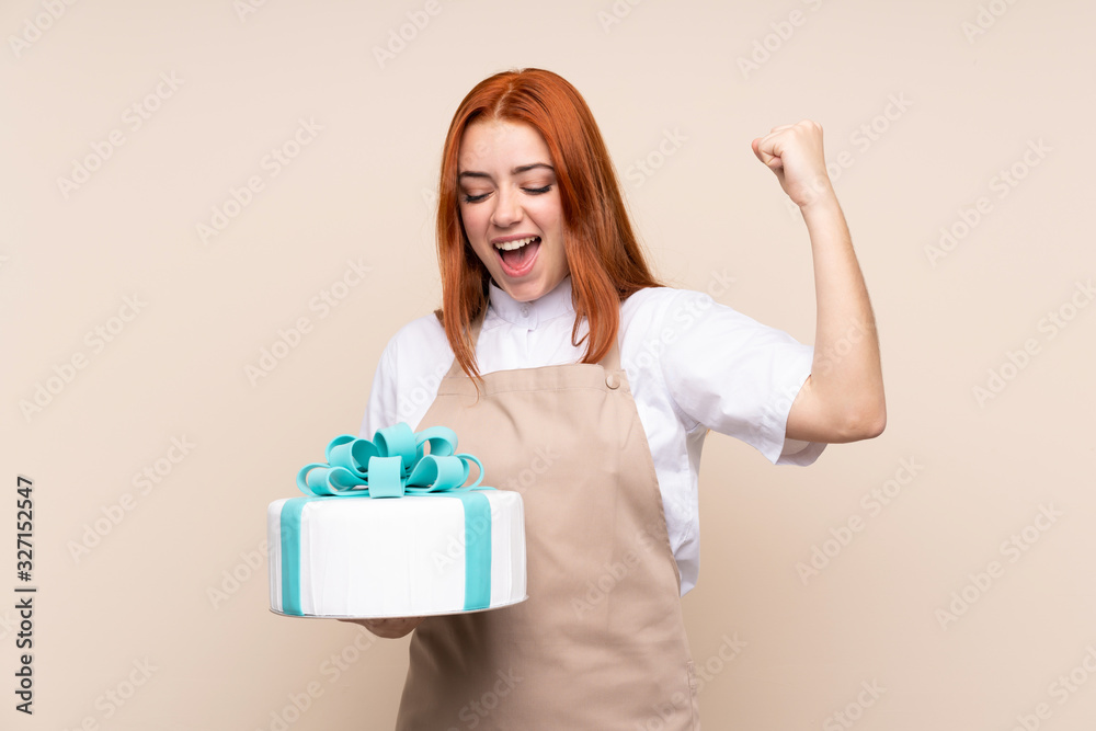 Redhead teenager girl with a big cake over isolated background celebrating a victory Redhead teenager girl with a big cake over isolated background celebrating a victory