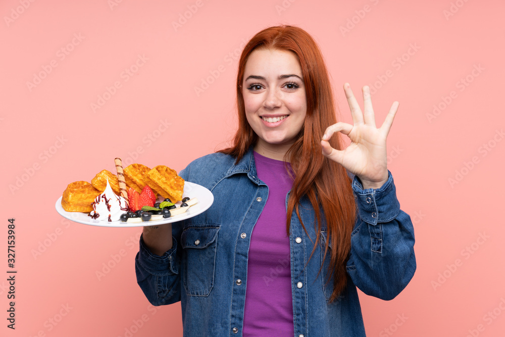 Redhead teenager girl holding waffles over isolated pink background showing ok sign with fingers Redhead teenager girl holding waffles over isolated pink background showing ok sign with fingers