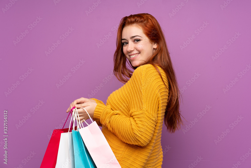 Redhead teenager girl over isolated purple background holding shopping bags and smiling Redhead teenager girl over isolated purple background holding shopping bags and smiling