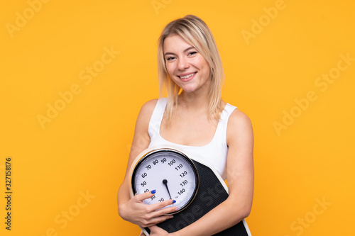 Young blonde Russian woman over isolated yellow background with weighing machine