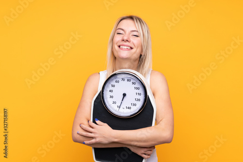 Young blonde Russian woman over isolated yellow background with weighing machine