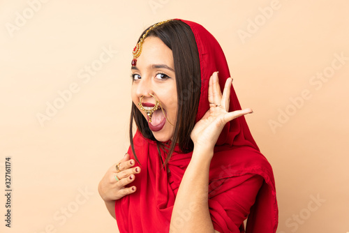 Young Indian woman isolated on beige background listening to something by putting hand on the ear © luismolinero
