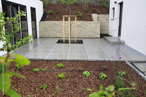 Moderner Gartenbau und Terrassengestaltung eines Neubaus an einem Hanggrundstück im Materialmix: Mauer aus Natursteinen, Pflastersteine aus Beton, Pflanzen und Bäume umgeben von Mulch und Schotter	