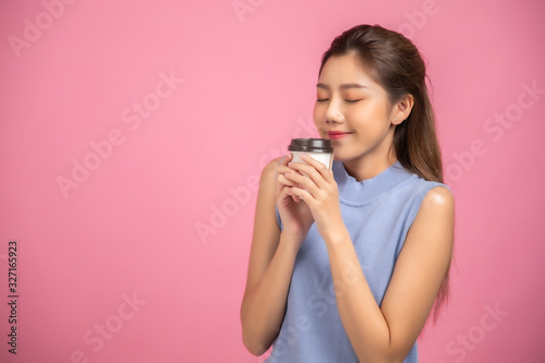 Portrait of a pretty young Asian business woman in sweater using mobile phone while holding takeaway coffee cup isolated over pink background.