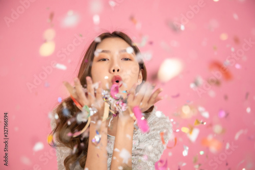 Portrait of a cheerful beautiful Asian womanl wearing dress standing standing under confetti rain and celebrating isolated over pink background.She blowing confetti from her hands .