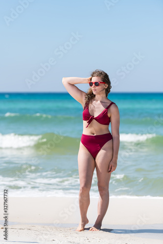 Young girl woman in red bikini bathing suit sunglasses touching hair with ocean sea green water in background in Santa Rosa Beach and horizon