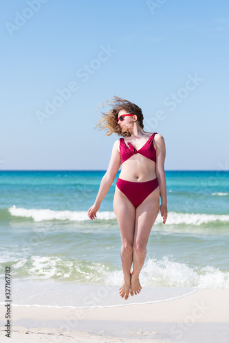 Young girl woman in red bikini bathing suit sunglasses jumping up with ocean sea green water in background in Santa Rosa Beach and horizon
