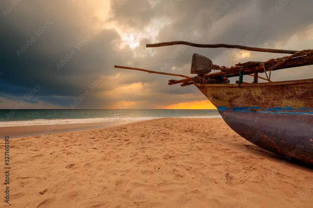 Fototapeta premium Traditional fisherman's boat on the sand of Sri Lanka ocean beach, under dramatic sunset sky in front of turquoise ocean waves.