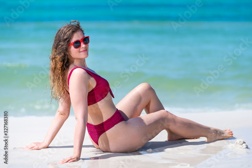 Young woman happy girl sitting down on sand in red swimsuit bikini bathing suit by ocean shore background in Florida