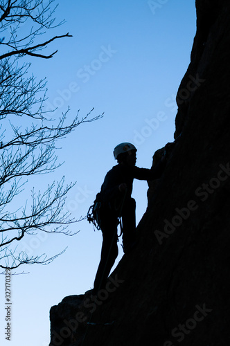 A dark silhouette of a rock climber climbing up a cliff on the background of blue sky and tree branches.