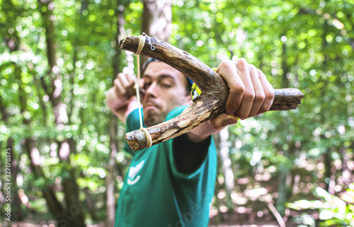 Canvas-taulu The man in the green shirt in the woods aims a large wooden slingshot in the camera lens