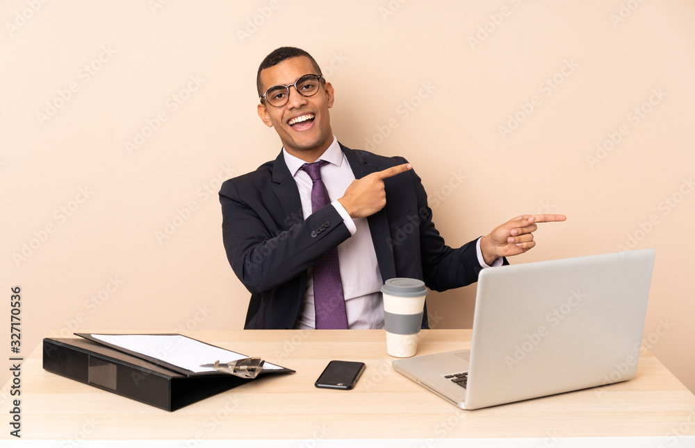 Young business man in his office with a laptop and other documents surprised and pointing side Young business man in his office with a laptop and other documents surprised and pointing side