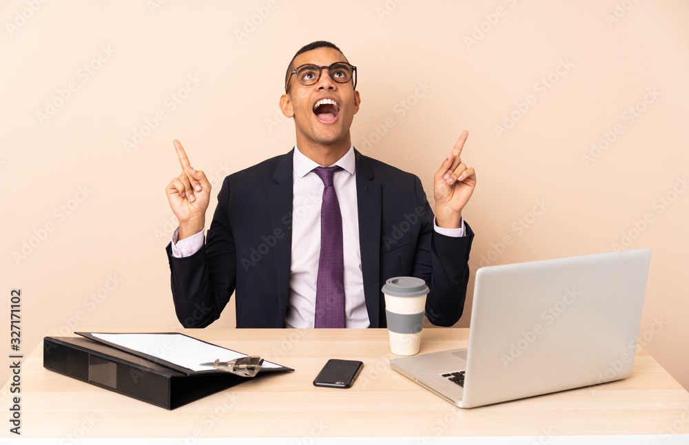 Young business man in his office with a laptop and other documents surprised and pointing up Young business man in his office with a laptop and other documents surprised and pointing up