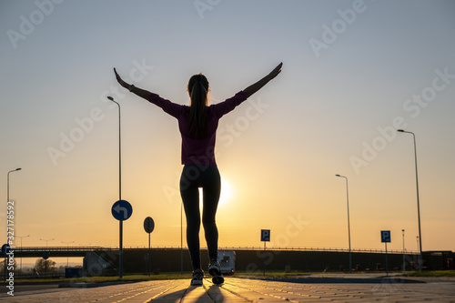 Dark silhouette of a young woman standing on empty parking lot at sunset.