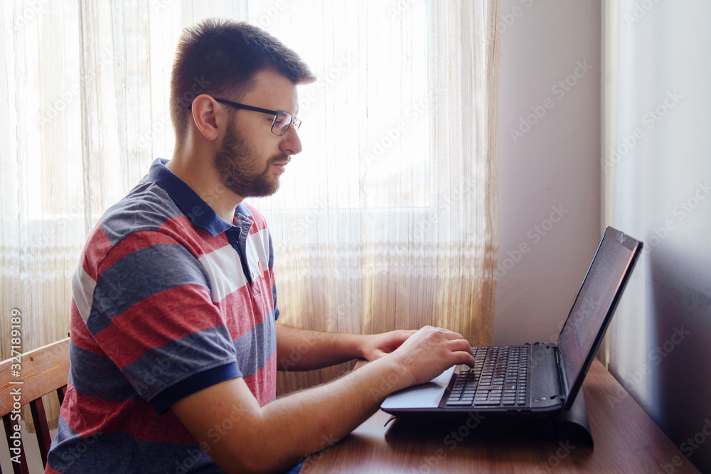 Young caucasian man adult wearing t-shirt and eyeglasses sitting by the table short hair and beard working on computer laptop at home browsing internet side view Young caucasian man adult wearing t-shirt and eyeglasses sitting by the table short hair and beard working on computer laptop at home browsing internet side view