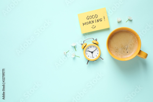 Delicious coffee, alarm clock and card with GOOD MORNING wish on light blue background, flat lay. Space for text