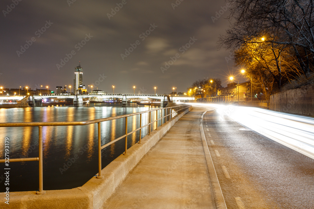 The lights of the city of Moscow and vehicles on the Krutitskaja embankment. An empty sidewalk at night.