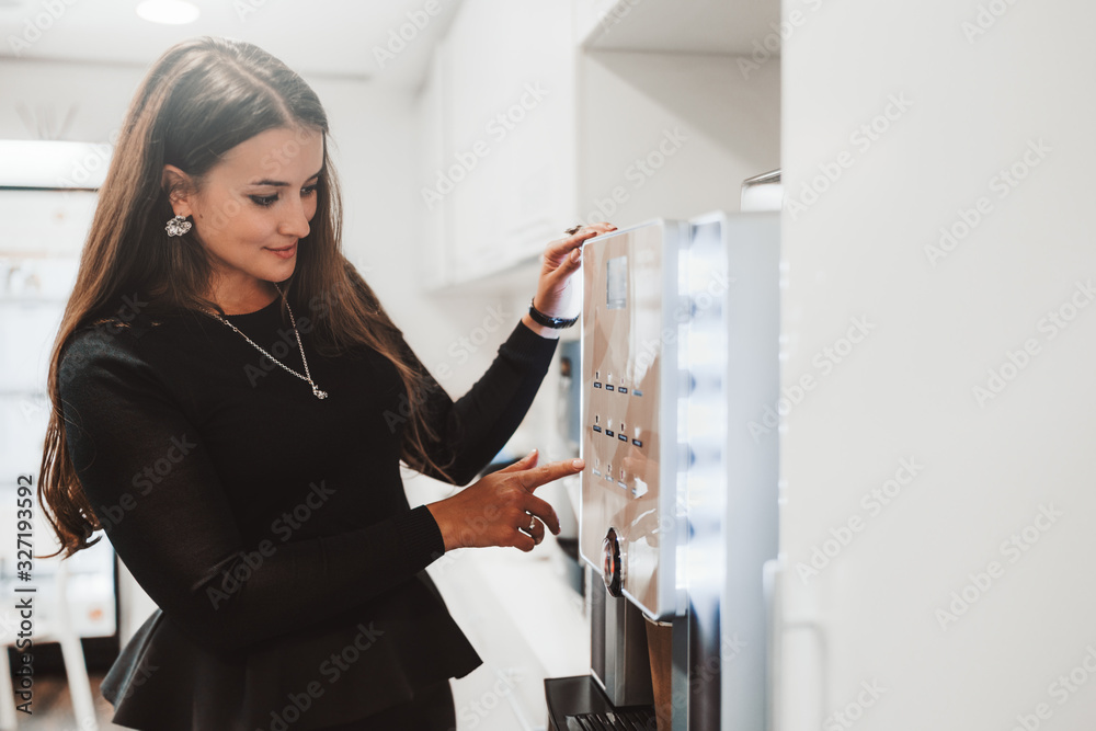 An adult caucasian woman entrepreneur is choosing a type of coffee to prepare while standing indoor of a cozy office coffee point on a kitchen in front of a modern coffee machine An adult caucasian woman entrepreneur is choosing a type of coffee to prepare while standing indoor of a cozy office coffee point on a kitchen in front of a modern coffee machine