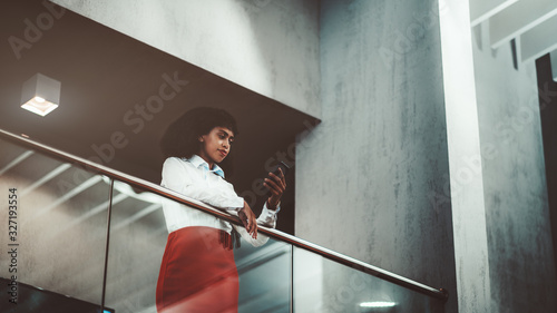 A pensive young African-American woman entrepreneur in a white shirt and red skirt is replying a message using her cellphone while leaning on chrome railing of a balcony in an office open-space area