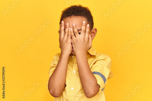 Frightened dark skinned little boy covering face with both hands being afraid of watching scary movie, peeping through hole between fingers. Confused timid child feeling shy hiding his emotions