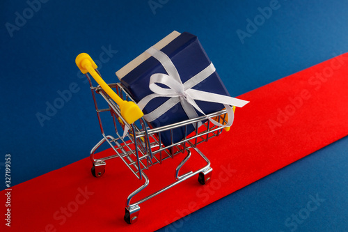 A gift with a white bow in a yellow shopping basket on the red carpet. Selective focus.