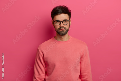 Waist up shot of serious male manager or freelancer looks at camera with calm expression, focused somewhere, comes on job interview, wears transparent glasses and sweater, poses over rosy background