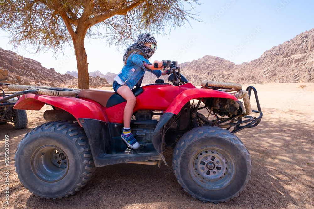 Brave kid in checkered scarf around head is riding red quad bike in sand desert Brave kid in checkered scarf around head is riding red quad bike in sand desert
