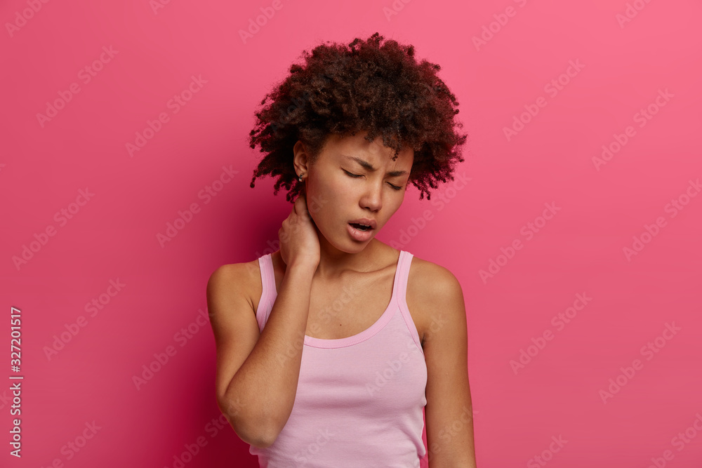 Photo of distressed woman touches neck, suffers from back or spine disease, yawns and closes eyes, has tired expression, dressed casually, has painful feelings, isolated over pink background Photo of distressed woman touches neck, suffers from back or spine disease, yawns and closes eyes, has tired expression, dressed casually, has painful feelings, isolated over pink background